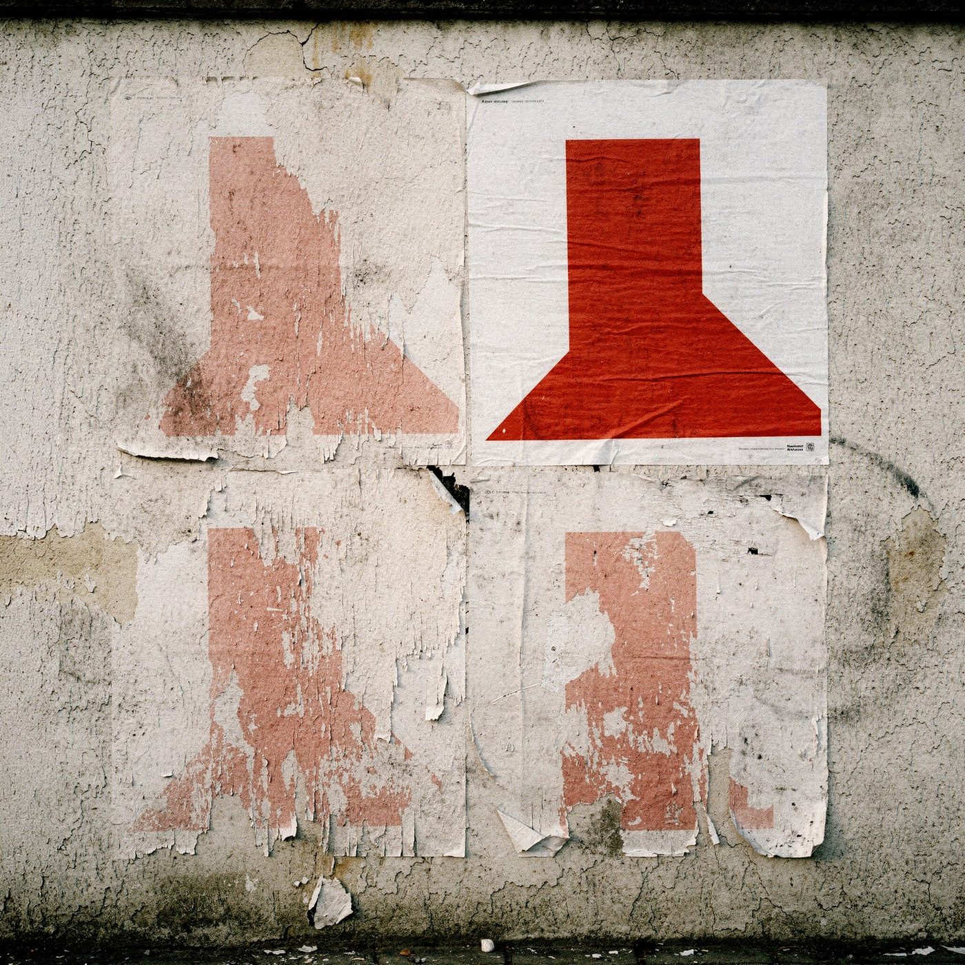 Quattro manifesti identici con un segno rosso affissi su un muro: tre consumati dal tempo, uno intatto